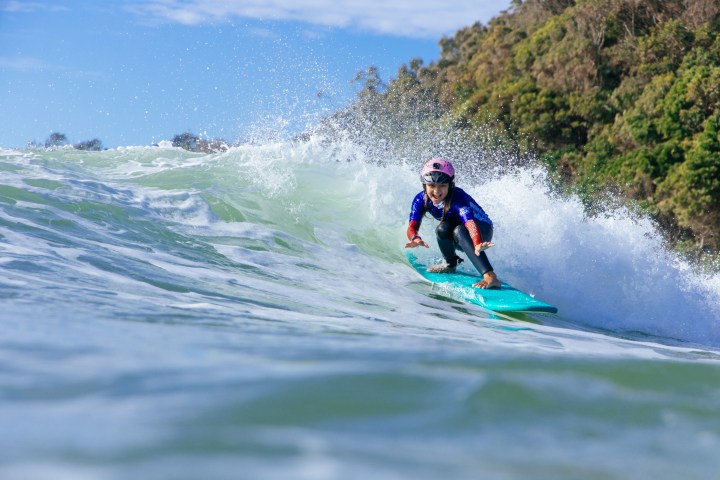 Person wearing a helmet surfing on a wave with a lush, green hill in the background.