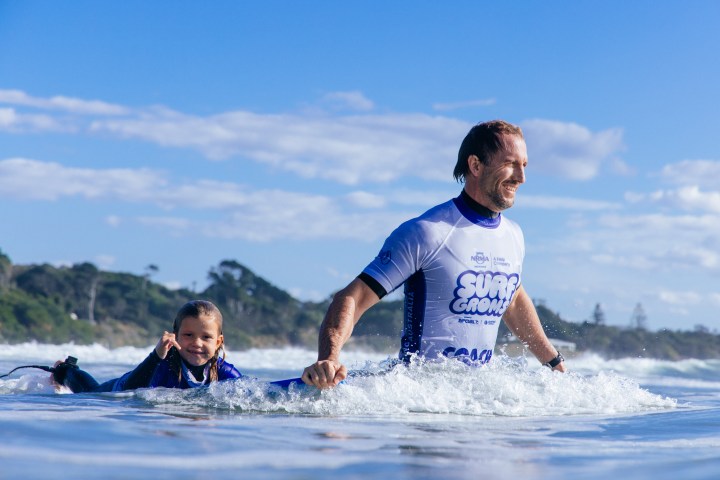 Child on surfboard and man in wetsuit in ocean, smiling, with waves and trees in background.