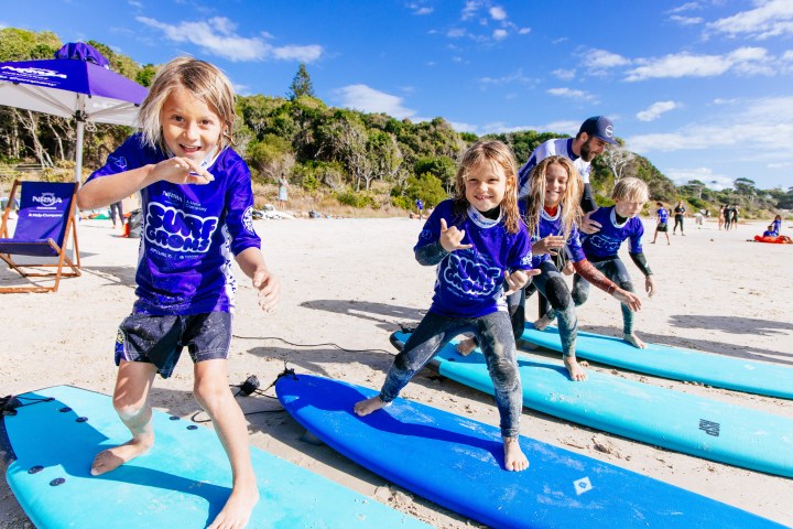 Children in blue shirts practicing surfing on the beach with blue surfboards.