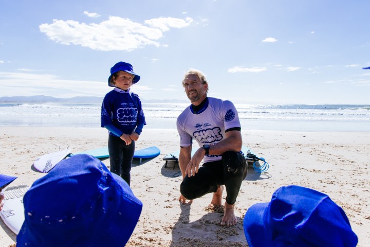 Instructor and child in blue surf gear on a sandy beach with surfboards and ocean in background.