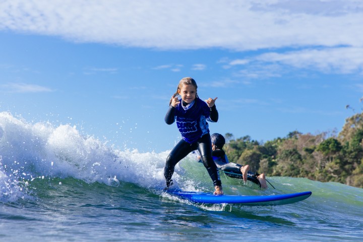 Two kids surfing a wave, one up front smiling, wearing blue shirts under a sunny sky.