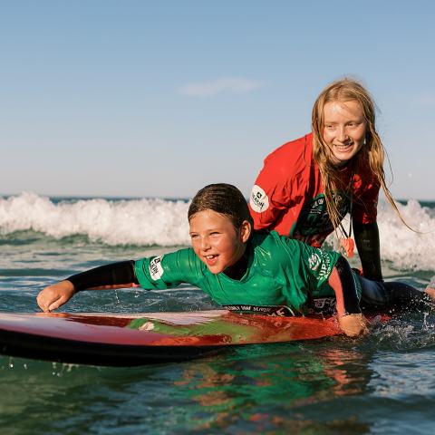 Two kids surfing, one on a board, in sunny ocean waves.
