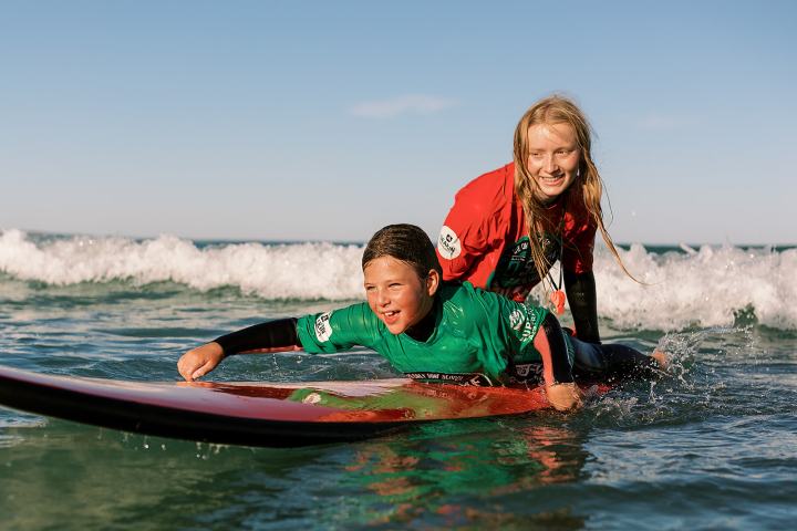 Two kids surfing, one on a board, in sunny ocean waves.