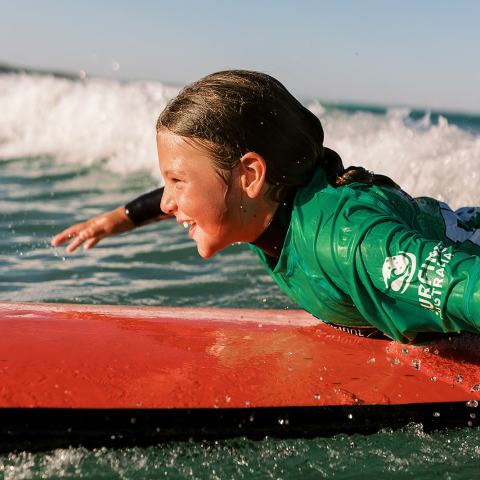 Child in green wetsuit on red surfboard smiling on ocean wave.