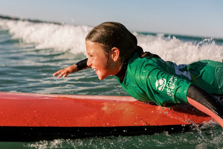 Child in green wetsuit on red surfboard smiling on ocean wave.