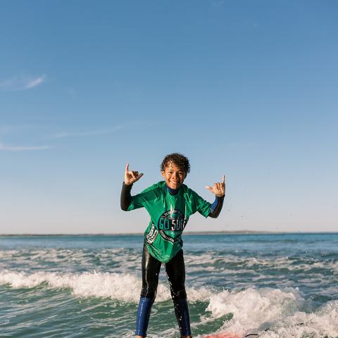 Person surfing on red board, wearing green shirt, making hand signs.
