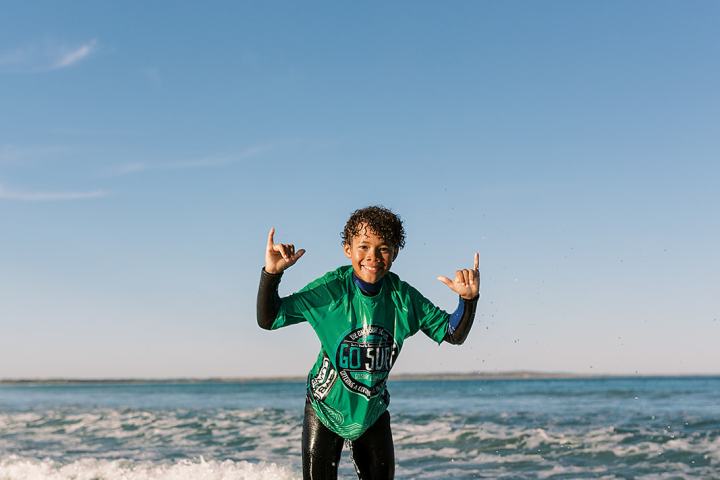 Person surfing on red board, wearing green shirt, making hand signs.
