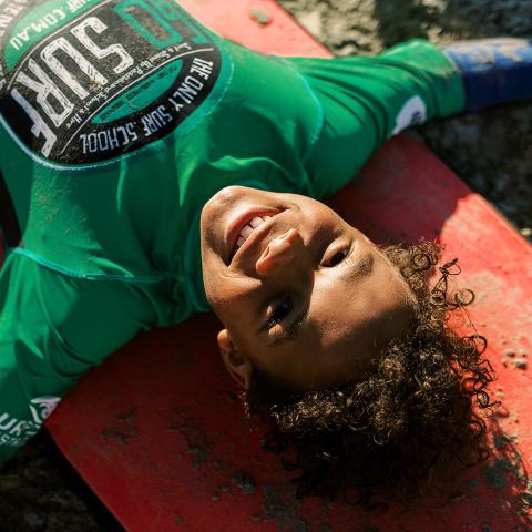 Child smiling, lying on a red surfboard on a sandy beach.