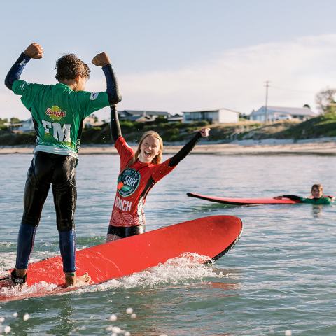 Two kids surfing on red boards, celebrating, with another surfer in the water.