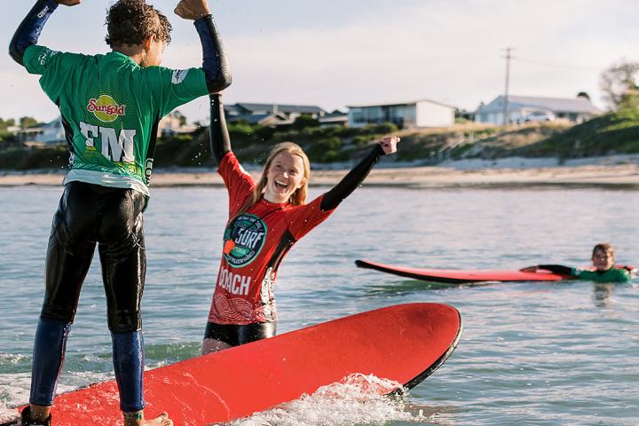 Two kids surfing on red boards, celebrating, with another surfer in the water.