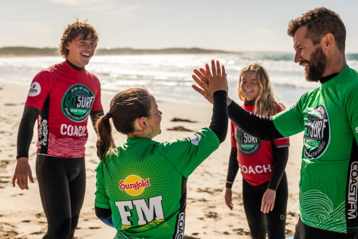 Surfers on beach high-fiving, wearing Go Surf jerseys, sunny day.